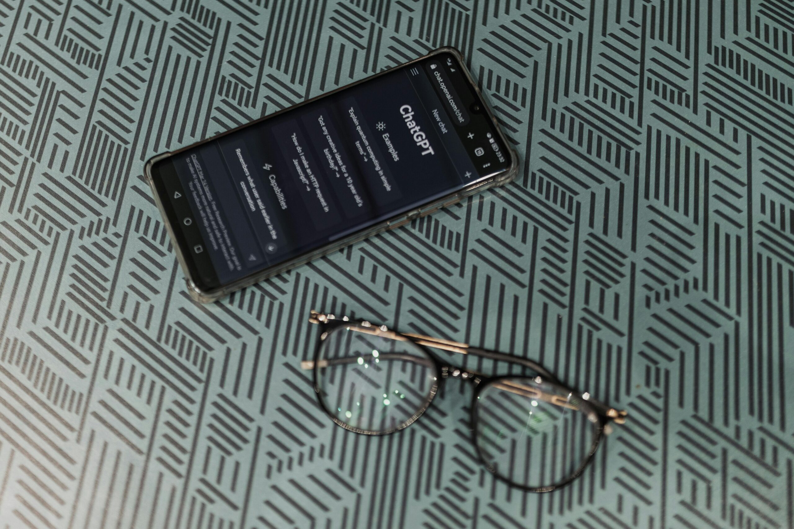 Close-up of smartphone displaying ChatGPT alongside eyeglasses on a patterned surface highlighting technology and style.